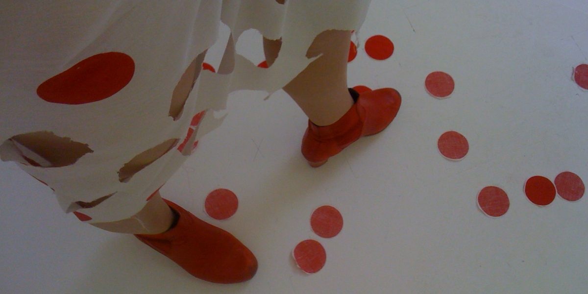 Eine Person in einem weißen Kleid mit roten Tupfen und roten Schuhen. Ein paar Tupfen sind auf dem Boden