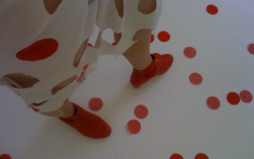 Eine Person in einem weißen Kleid mit roten Tupfen und roten Schuhen. Ein paar Tupfen sind auf dem Boden