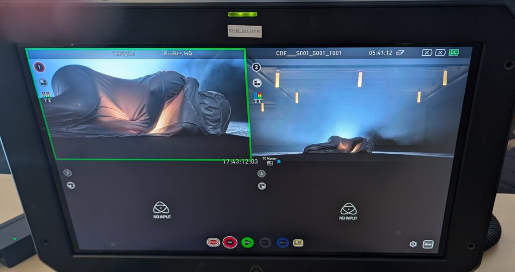 Monitor-Ansicht einer Aufnahme, eine Figur in einer silbernen Hülle liegt am Boden.
